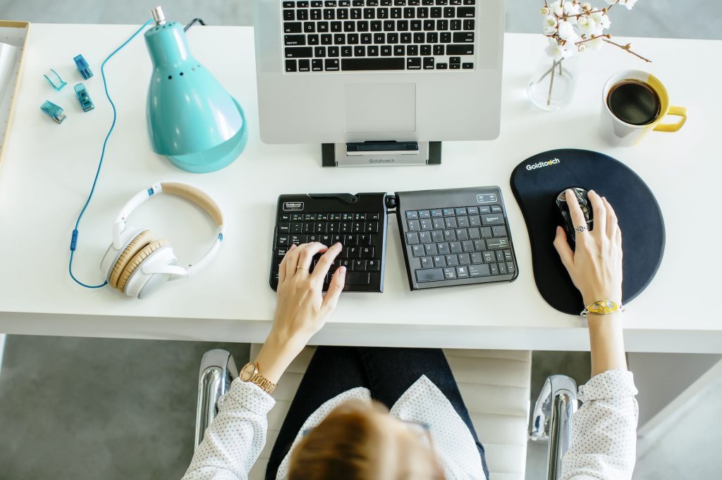 Woman sitting at an ergonomically positioned workstation. Woman is keying the keyboard, mousing the mouse, and looking at a laptop positioned on a Goldtouch laptop stand