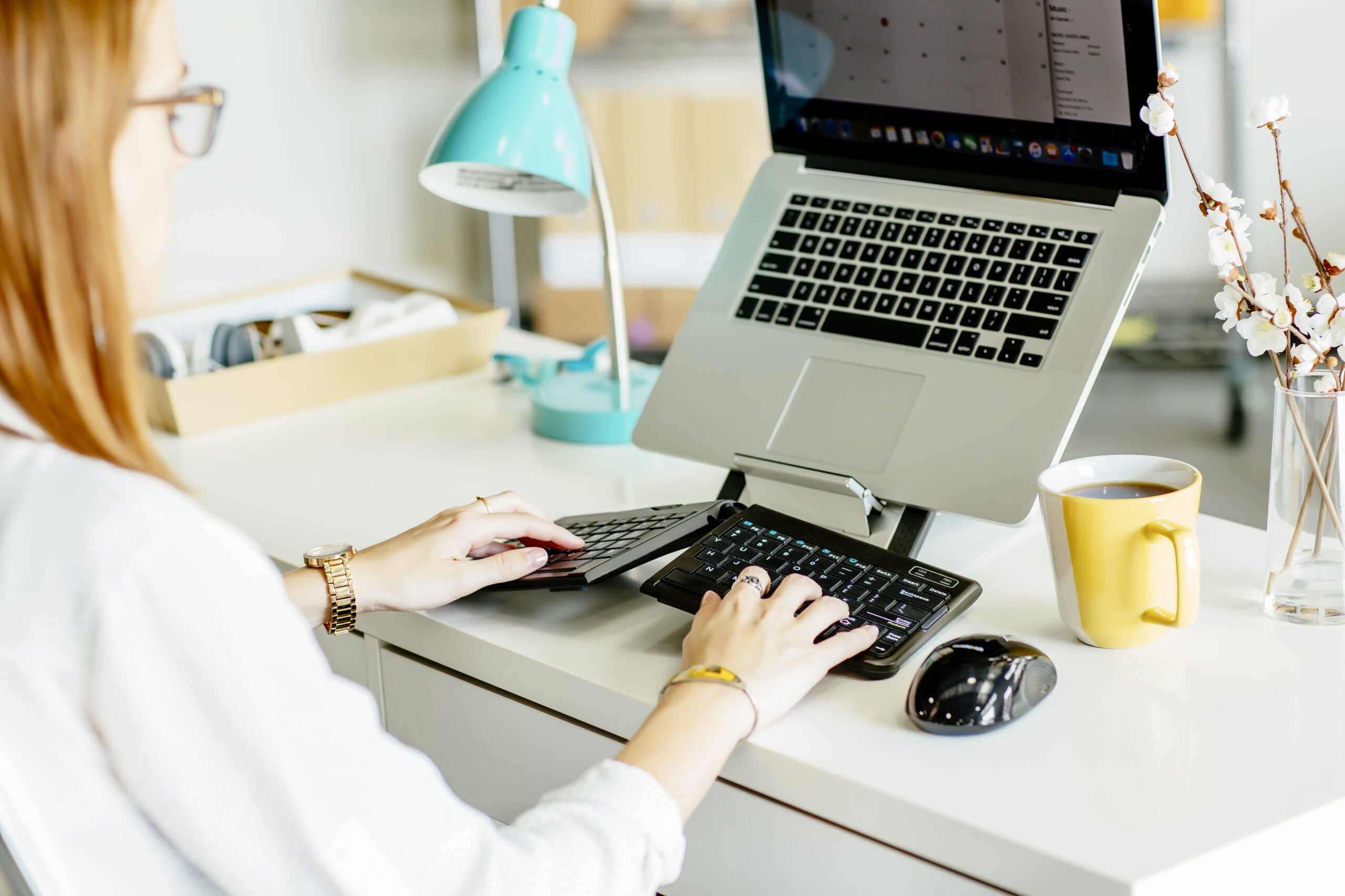 Woman sitting at a desk with her hands typing on a Goldtouch Go!2 Mobile Keyboard. She is looking at a laptop that is on a Goldtouch Laptop/Tablet Stand in front of her. There is a Goldtouch Ambi Mouse next to her keyboard ready for use. 