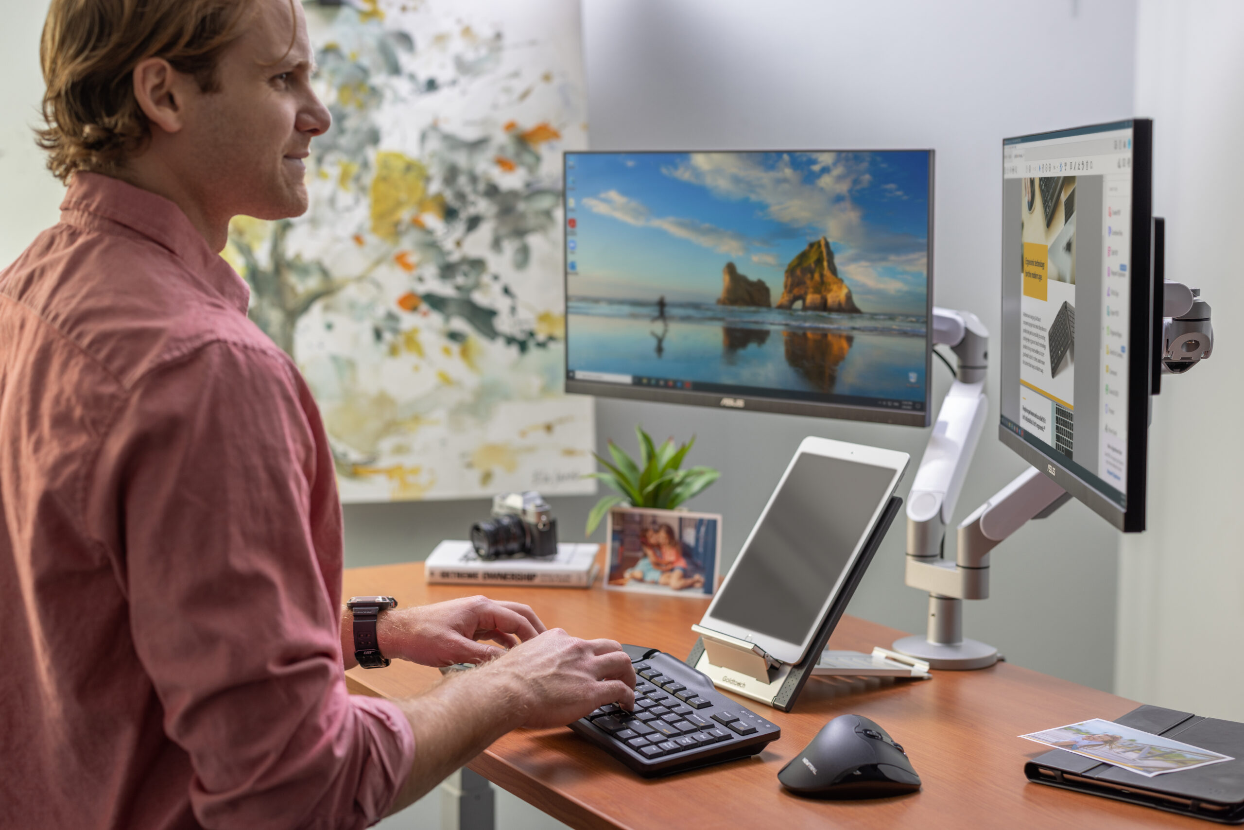 Elite Keyboard for Ultimate Comfort. A lifestyle image of a man at his ergonomic desktop featuring a Goldtouch Elite Keyboard, Newtral Mouse, Laptop Stand with an iPad on it, Monitor Arms, various desktop accessories like pictures, a book, camera, decorative plant, and notebook.