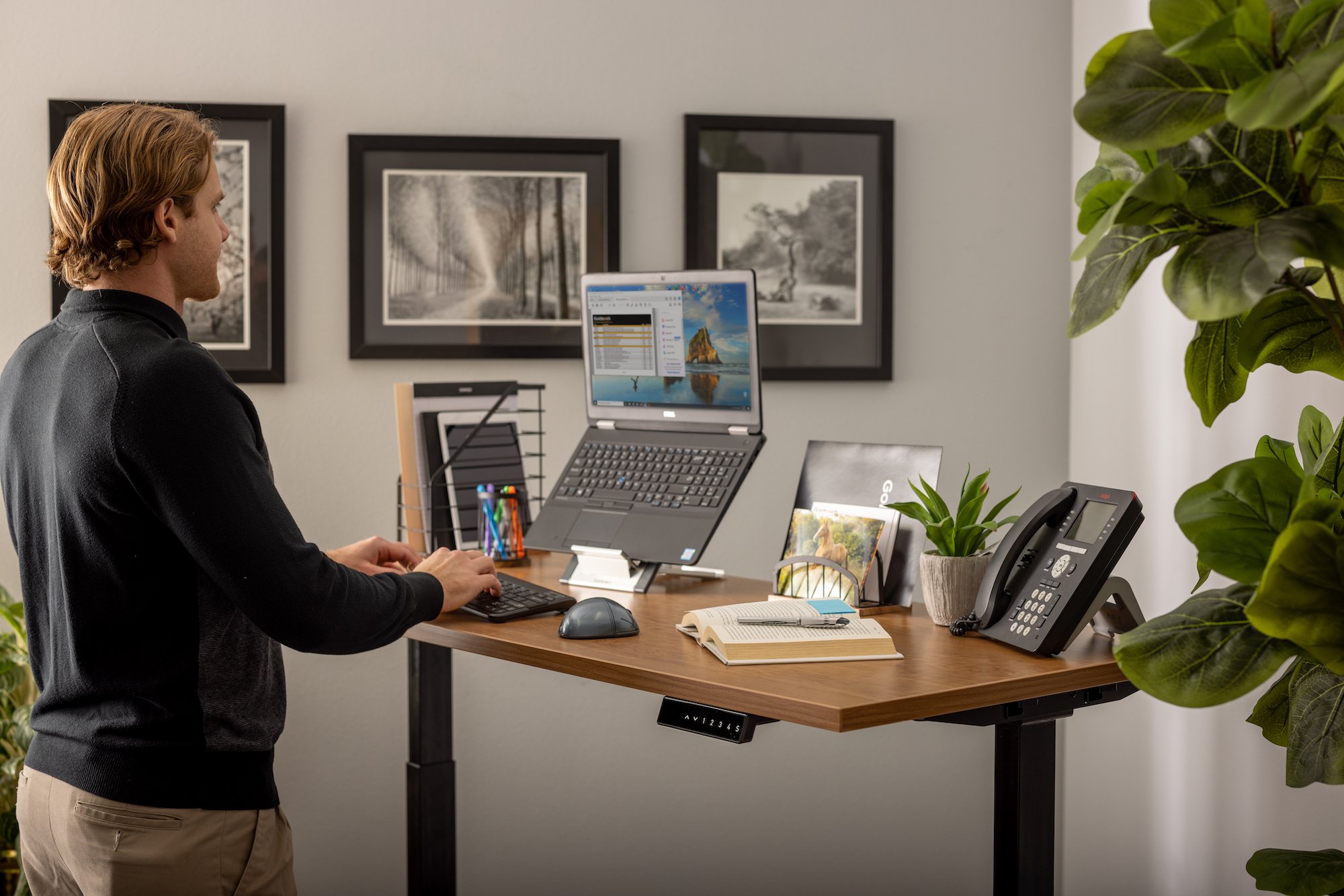 A lifestyle image of a man standing at his desk typing on a Goldtouch Go!2 Mobile Keyboard and a FlexMouse next to it. There's a Laptop Stand with a laptop on it, a phone, plant, book, and other desktop accessories.
